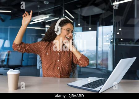 Junge, schöne hispanische Frau mit Brille und schwarzen Lockenhaaren, die im Büro arbeitet, Geschäftsfrau, die sich ausruht und Musik in Kopfhörern hört, die in der Pause tanzen und singen Stockfoto