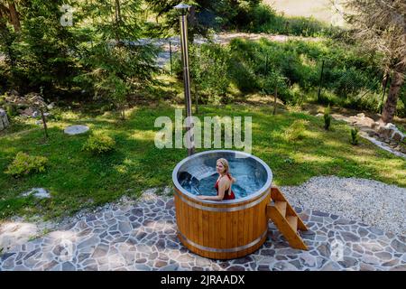 Junge Frau, die in der Holzbadewanne mit Kamin Holz verbrennt und Wasser im Hinterhof in den Bergen erwärmt. High-Angle-Ansicht. Stockfoto