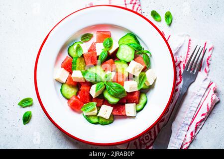 Wassermelonensalat mit Fetakäse, Gurke und Basilikum auf einem Teller, grauer Hintergrund, Draufsicht. Sommerrezept, gesunde Ernährung, Detox-Lebensmittel. Stockfoto