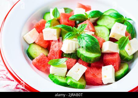 Wassermelonensalat mit Fetakäse, Gurke und Basilikum auf einem Teller, grauer Hintergrund, Nahaufnahme. Sommerrezept, gesunde Ernährung, Detox-Lebensmittel. Stockfoto