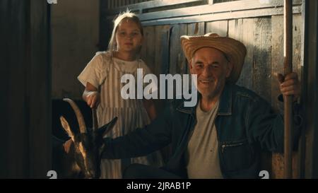 Glücklicher älterer Mann mit lächelndem Werkzeug und Blick auf die Kamera, während er in der Nähe von niedlichem Mädchen und Ziege in der Scheune auf der Ranch sitzt Stockfoto