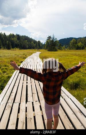Kaukasisches Mädchen, Rückenansicht, mit offenen Armen in einer natürlichen Berglandschaft auf einem Holzweg. Naturpark Pian di Gembro, Valtellina, Italien. Stockfoto
