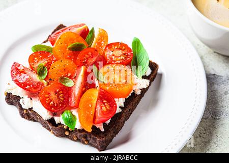 Roggen-Toast mit Frischkäse, Tomaten und Basilikum auf weißem Teller und einer Tasse Kaffee. Gesundes Frühstücksrezept. Stockfoto