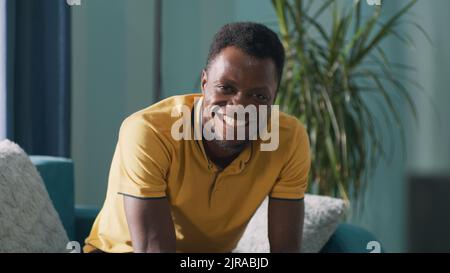 Ein glücklicher afroamerikanischer Mann, der mit einem Lächeln und klirrenden Fäusten auf die Kamera blickt, während er auf dem Sofa sitzt und zu Hause den Sieg feiert Stockfoto