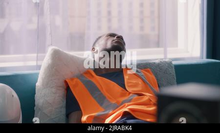 Erschöpfter schwarzer Mann in der Weste, der aufwacht und einen Hardhut aufsetzt, dann vom Sofa aufsteht und zur Arbeit geht Stockfoto