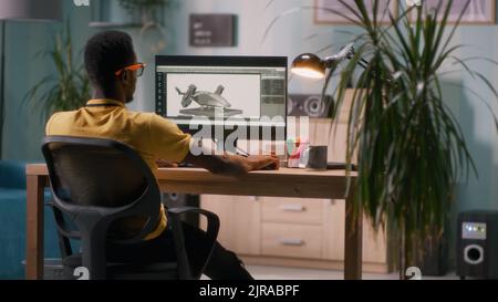 Afroamerikanischer Mann, der 3D Modell und Video für Videospiele am Computer erstellt, während er am Tisch in einem hellen Arbeitsplatz zu Hause sitzt Stockfoto