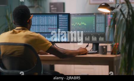 Rückansicht eines afroamerikanischen Mannes, der Daten auf Computern analysiert, während er Krypto-Währung während der Fernarbeit von zu Hause aus handelte Stockfoto
