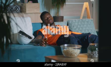 Erschöpfter schwarzer Mann in orangefarbener Weste trinkt Bier und schaut sich einen Film im Fernsehen an, während er sich abends auf der Couch entspannt Stockfoto