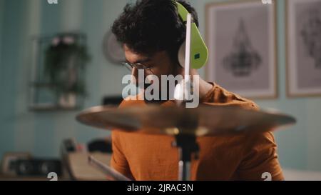 Bärtiger indischer Schlagzeuger in kabellosen Kopfhörern, der während der Probe im Heimstudio rhythmische Melodie auf Trommeln spielt Stockfoto