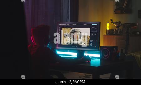 Männlicher Teenager, der Computer verwendet, um das Bild des Astronauten zu retuschieren, während er am Tisch sitzt und nachts zu Hause die Bearbeitungssoftware lernt Stockfoto