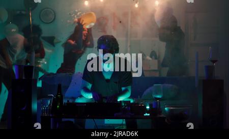 Zoomen Sie mit dem bärtigen Mann in einer stilvollen Sonnenbrille, der die Kamera anschaut, während er auf der Couch sitzt und während einer wilden Party in der Nacht zu Hause elektronische Musik spielt Stockfoto