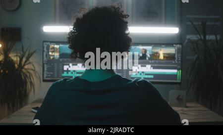 Nicht erkennbarer Typ mit lockigen Haaren Bearbeitung Video auf dem Computer unter heller Lampe, während fernab von zu Hause in dunklen Raum in der Nacht arbeiten Stockfoto