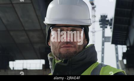 Mann mit Schutzbrille und Hardhut, der auf der Baustelle mit ernstem Gesicht gegen Betonbrücken-Stützen auf die Kamera schaut Stockfoto