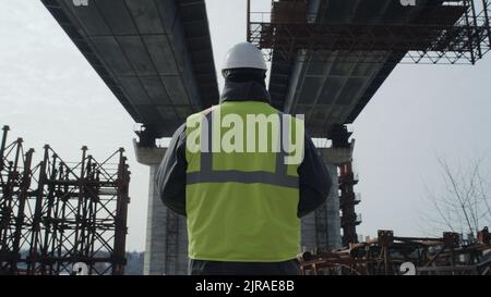 Ansicht eines anonymen Mannes in Uniform und Harthut, der die unfertige Betonbrücke auf der Baustelle inspiziert Stockfoto