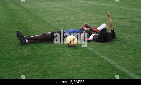 Schwenken Sie um die Ansicht eines schwarzen Mannes, der während der Trainingspause auf dem Fußballfeld liegt und einen gemischten Rennjungen kitzelt und in den Himmel zeigt Stockfoto