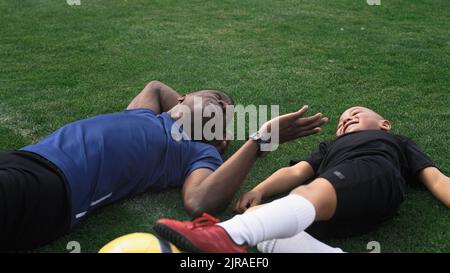 Schwenken Sie um die Ansicht eines schwarzen Mannes, der während der Trainingspause auf dem Fußballfeld liegt und einen gemischten Rennjungen kitzelt und in den Himmel zeigt Stockfoto