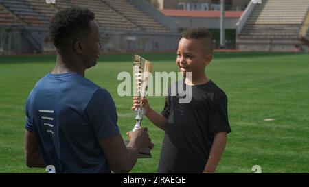 Afroamerikanischer Mann, der nach einem Fußballspiel im Stadion Trophäe übergab und mit einem gemischten Rennjungen sprach Stockfoto