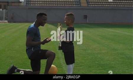 Afroamerikanischer Mann, der nach einem Fußballspiel im Stadion Trophäe übergab und mit einem gemischten Rennjungen sprach Stockfoto