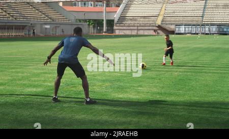Afroamerikanischer Mann, der versucht, den Ball zu fangen, der von einem gemischten Rennjungen geschossen wurde, während er im Sommer auf dem grünen Feld im Stadion Fußball spielt Stockfoto