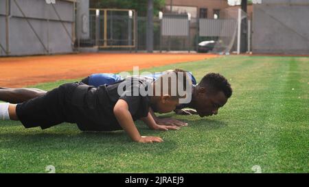 Schwenken Sie um die Ansicht eines schwarzen Trainers und eines gemischten Rennjungen in Sportkleidung, der während des Fußballtrainings im Stadion Liegestütze auf dem Spielfeld macht Stockfoto