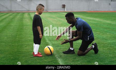 Schwenken Sie um die Ansicht eines afroamerikanischen Mannes, der einem Jungen mit gemischtem Rennen erklärt, wie er während des Fußballtrainings auf dem Spielfeld mit dem Ball umgeht Stockfoto