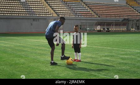 Schwenken Sie um die Ansicht eines Kindes mit gemischtem Rennen, das um einen afroamerikanischen Trainer herumläuft und versucht, während des Fußballtrainings auf dem Spielfeld Ball zu nehmen Stockfoto