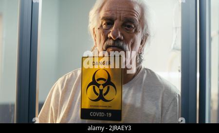 Gealterter Mann, der im modernen Krankenhaus hinter einer Glaswand der Coronavirus-Station stand und wegschaute Stockfoto