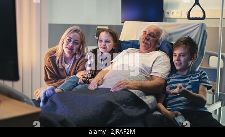 Ein älterer Mann lächelt und redet mit Tochter und Enkeln, während er auf der Krankenstation im Bett liegt und fernsieht Stockfoto