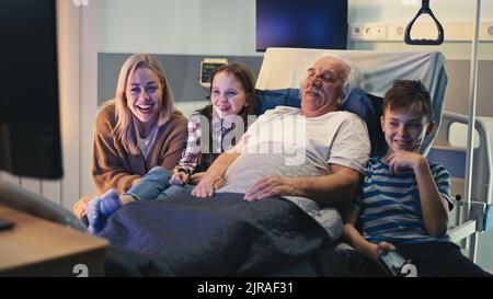 Ein älterer Mann lächelt und redet mit Tochter und Enkeln, während er auf der Krankenstation im Bett liegt und fernsieht Stockfoto