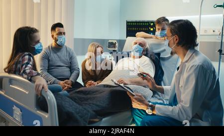 Arzt mittleren Alters in Maske im Gespräch mit älteren männlichen Patienten und Familie auf der Station der modernen Klinik während der Pandemie Stockfoto