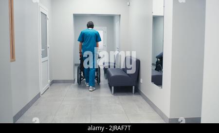 Rückansicht eines Mannes in einem Peelings, der mit einem behinderten männlichen Patienten den Rollstuhl schiebt, während er im Korridor einer modernen Klinik arbeitet Stockfoto