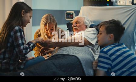 Älterer Mann, der auf der Krankenstation auf dem Bett liegt, spricht und der glücklichen erwachsenen Frau und Kindern das Pulsoximeter zeigt Stockfoto