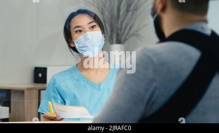 Asiatische Frau in Maske, die einem männlichen Patienten mit gebrochenem Arm Papier gab und bei der Arbeit an der Rezeption in einer modernen Klinik Richtung zeigte Stockfoto