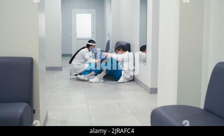 Frau mit Gesichtsschutz und Maske, die während einer Pandemie Platz nimmt und einen erschöpften Arzt mittleren Alters auf dem Flur eines modernen medizinischen Zentrums unterstützt Stockfoto
