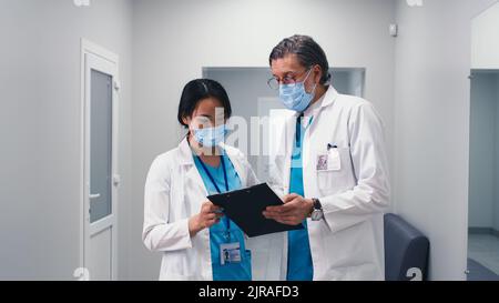 Reifer Mann in Maske diskutiert Daten auf Zwischenablage mit asiatischen weiblichen Kollegen dann zu Fuß weg während der Arbeit im medizinischen Zentrum Stockfoto