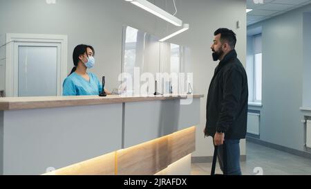 Bärtiger Mann in legerer Kleidung, der mit einer weiblichen asiatischen Rezeptionistin in Maske und einem reifen männlichen Arzt spricht, während er ein modernes Krankenhaus besucht Stockfoto