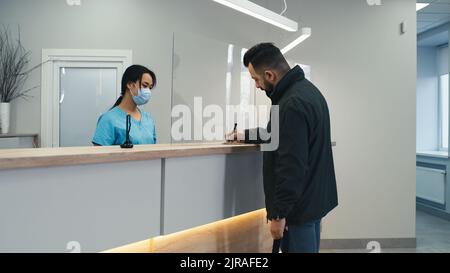 Bärtiger Mann, der mit einer asiatischen Rezeptionistin in Maske sprach und den Vertrag unterzeichnete, während er das moderne medizinische Zentrum besuchte Stockfoto
