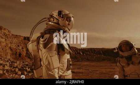 Mann im Raumanzug, der die Kamera auf dem Helm der sprechenden Frau justiert und den Daumen während der Erkundung des Bergrückens auf dem Mars hochstigelt Stockfoto