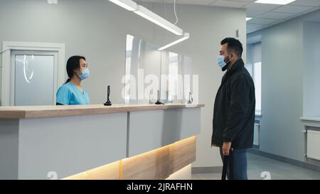 Asiatische Frau in Maske sprechen mit Patienten und nicken dann Papier zu reifen Arzt und warten auf Männer während der Arbeit zu verlassen Stockfoto