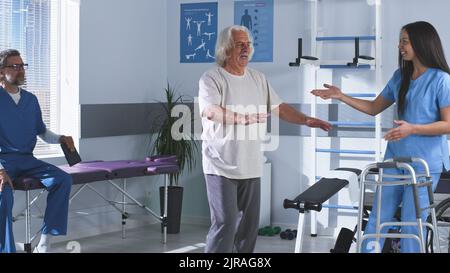 Erwachsene Frau, die älteren männlichen Patienten hilft, ohne Gehrahmen zu spazieren, während ein reifer Kollege eine Tablette verwendet, dann klatschen die Ärzte während der Rehabilitationssitzung im Krankenhaus mit den Händen Stockfoto