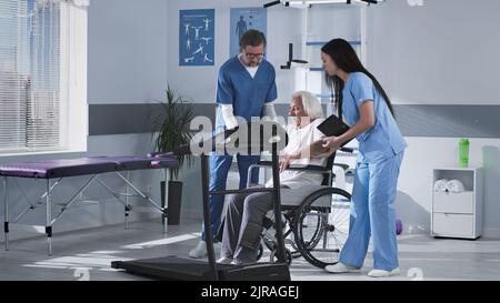 Ärzte helfen älteren Menschen, auf dem Laufband zu klettern und erklären Rehabilitationsübungen während der Arbeit in einer modernen Klinik Stockfoto