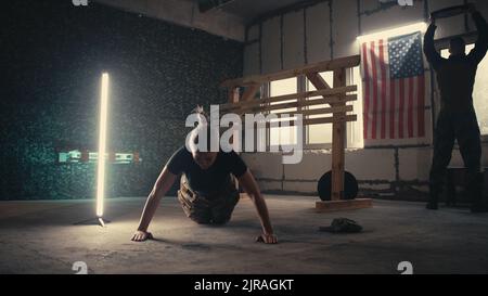 Ansicht der Frau, die Liegestütze in der Nähe des Mannes macht, der während des intensiven Trainings im Fitnessstudio des US-Militärstützpunktes mit Gewicht trainiert Stockfoto