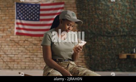 Afroamerikanische Militärfrau, die auf der Bank sitzt und mit der USA-Flagge auf der Basis auf dem Smartphone gegen eine Backsteinmauer surft Stockfoto