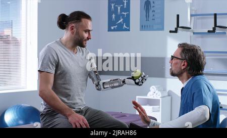 Ein glücklicher Mann, der den Tennisball im bionischen Arm ergreift und bewegt, während er einen reifen Arzt in einer modernen Klinik besucht Stockfoto