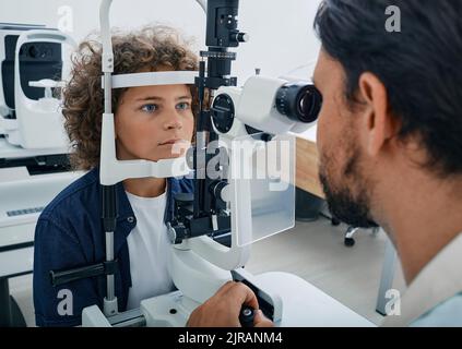Lockiger Junge, der Augenuntersuchung in der Augenklinik mit Optiker erhält. Kontrolle der Netzhaut des männlichen Kinderauges, Augenheilkunde für Kinder Stockfoto