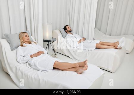 Ein Paar während des romantischen Wochenendes. Ein liebes junges Paar in weißen Bademänteln, das sich im Wellnessbereich auf weißen Betten in der Lounge-Zone niederlegt Stockfoto