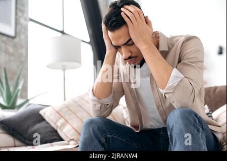 Frustrierter, trauriger indischer oder arabischer Mann, sitzt zu Hause auf dem Sofa im Wohnzimmer mit geschlossenen Augen, hält die Hände am Kopf, leidet unter Kopfschmerzen, erlebt persönliche Probleme. Kopfschmerzen, Migräne Stockfoto