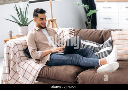 Positiver arabischer oder indischer junger Mann in lässiger, stilvoller Kleidung, Freiberufler, Webdesigner, sitzt auf dem Sofa im Wohnzimmer, nutzt Laptop für die Arbeit an einem Projekt, elearning, Surfen im Internet, Lächeln Stockfoto