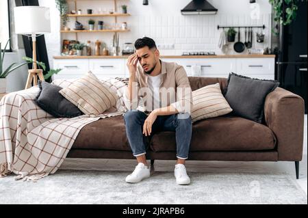 Einsamkeit. Frustriert besorgt verwirrt verärgert indischen oder arabischen jungen Mann in Freizeitkleidung, sitzt allein auf dem Sofa zu Hause im Wohnzimmer, erlebt persönliche Probleme, Sorgen, Depressionen, Stress Stockfoto