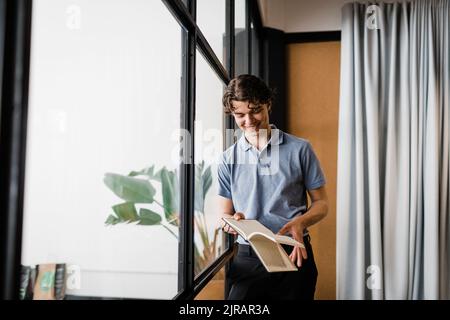 Lächelnder junger Geschäftsmann mit Tagebuch, das an einer Glaswand gelehnt ist Stockfoto